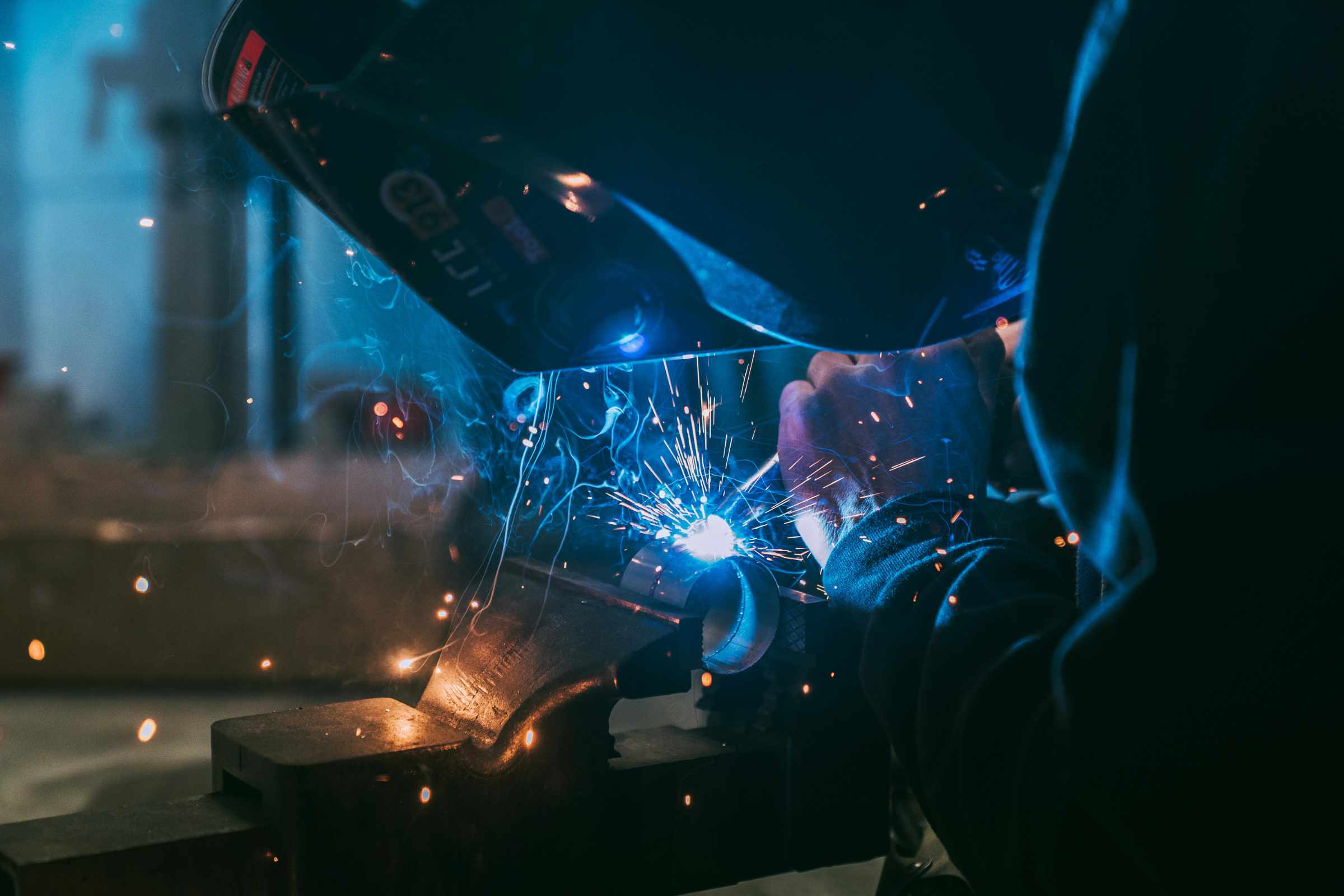 A man wearing a welding helmet is focused on welding a metal piece on a work table.