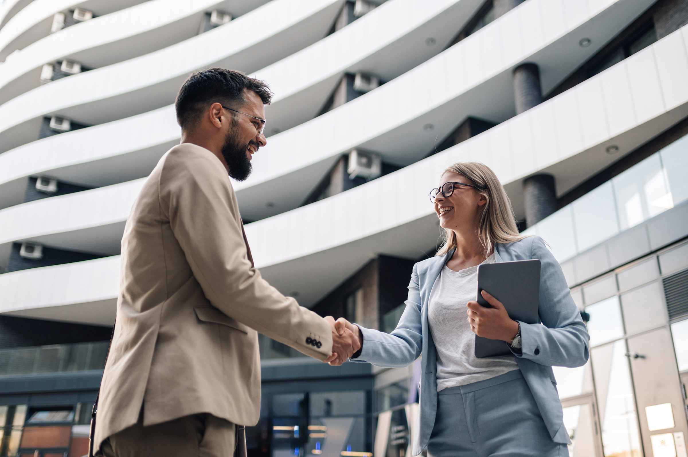 Two smiling business professionals shake hands outside a modern office building with curved white balconies